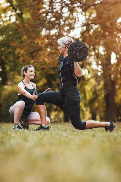 A muscular mature woman is doing exercises with personal trainer in the park, dressed in a black suit with an EMS electronic simulator to stimulate her muscles.