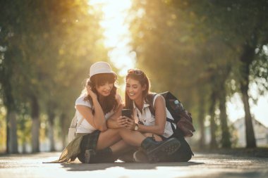 Sırtlarında sırt çantaları olan iki güzel genç kadın akıllı bir telefonla sosyal medyada geziniyorlar ve sonbahar güneşli caddede oturuyorlar..