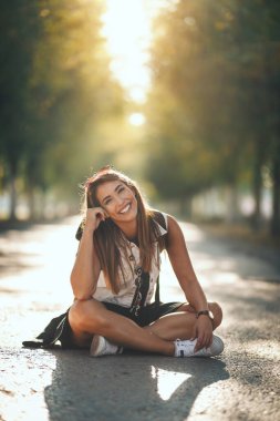 Sırtında sırt çantaları olan genç ve güzel bir kadın sonbahar güneşli caddede oturuyor ve kameraya bakıyor..