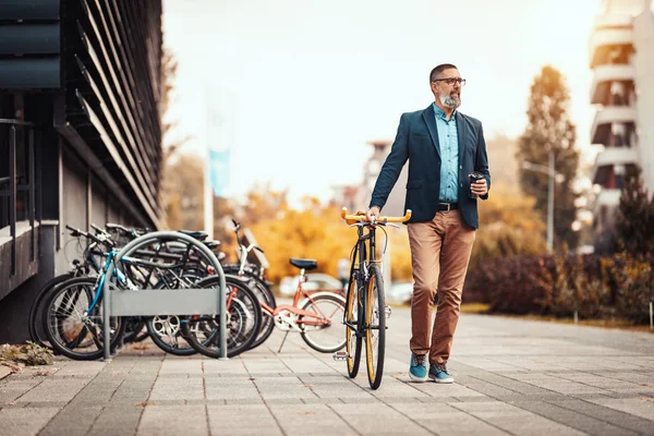 コーヒーを飲みながら自転車でオフィス街の前を歩くことに成功したハンサムな中年ビジネスマン — ストック写真