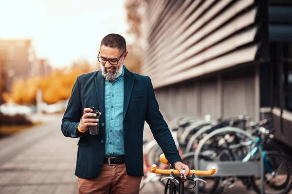 コーヒーを飲みながら自転車でオフィス街の前で仕事や散歩に行く成功したハンサムな中年のビジネスマン — ストック写真