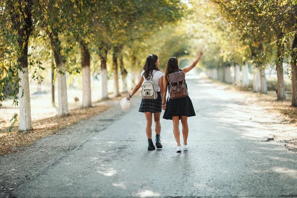 两个背着背包的年轻貌美的女人手牵着手 望着远方 沿着秋天阳光灿烂的大街走着 — 图库照片