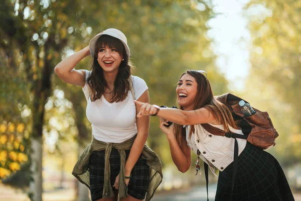 两个年轻美丽的微笑的女人背着背包 沿着秋天阳光灿烂的大街走着 互相交谈着 指点着什么 — 图库照片