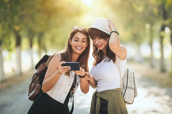 バックパックを背負った2人の美しい若い笑顔の女性が 秋の晴れた日の街を歩いているとスマートフォンの写真を見ている — ストック写真