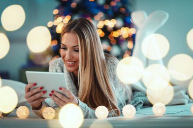 Tablet kullanan ve mutlu bir şekilde gülümseyen sevimli genç bir kadın evde rahat bir Noel tatili geçiriyor..