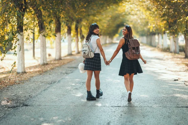 两个背着背包的年轻貌美的女人手牵手 望着相机 沿着秋天阳光灿烂的大街走着 — 图库照片