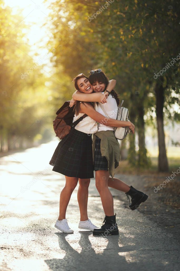 Two beautiful young smiling women with backpacks on their back are walking along the autumn sunny avenue and hugging.