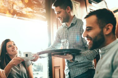 Genç ve yakışıklı bir garson bir kafede gülümseyen bir grup arkadaşa servis yapıyor..