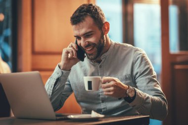 Genç ve mutlu bir adam kafede telefon görüşmesi yapıyor. Müşterilerle konuşuyor, laptopa bakıyor ve kahve içiyor..