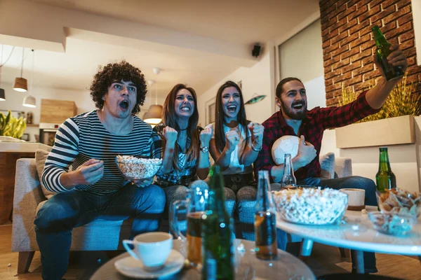 A group of friends watches the broadcast of a sports event. They sit in front of the TV in the living room, eat snack, drink beer, and cheer for the favorite team
