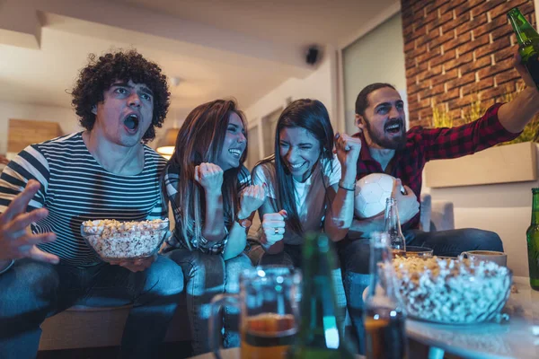 A group of friends watches the broadcast of a sports event. They sit in front of the TV in the living room, eat snack, drink beer, and cheer for the favorite team