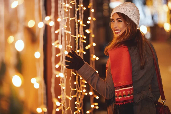 若い美しい笑顔の女性がお店の窓から何かを探しているクリスマスの時に街の通りで楽しんでいます — ストック写真