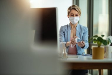 Tıbbi koruyucu maskeli bir iş kadını el dezenfeksiyonu için antibakteriyel jel kullanıyor ve grip salgını ve koronavirüs salgını sırasında enfeksiyonu önlemek için kendini izolasyon ve karantina sırasında bilgisayarın başında çalışıyor. .