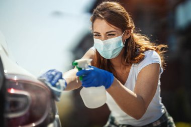 Yüzünde maske ve ellerinde koruyucu eldivenler olan genç bir kadın, birinin içinde bez, diğerinde dezenfektan olan bir şişe tutarak arabasını siler..