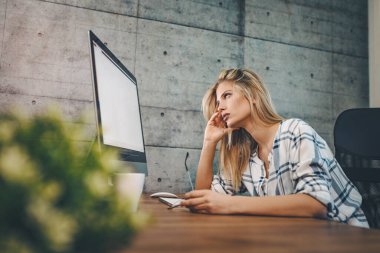 Yorgun ve yorgun genç bayan tasarımcı ofiste oturuyor. Bir bilgisayar başında ve çalışma saatlerinin bitmesini bekliyor..