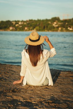 Gün batımında nehir kıyısında dinlenen hasır şapkalı genç bir kadının arkası..