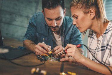 İki genç mühendis demir lehimleyerek elektronik ekipmanların onarımına odaklanmış durumdalar. Adam, bayanlara devre kartının elementlerini lehimlemede yardım ediyor.. 