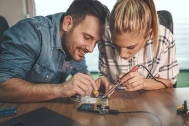 İki genç mühendis demir lehimleyerek elektronik ekipmanların onarımına odaklanmış durumdalar. Adam, bayanlara devre kartının elementlerini lehimlemede yardım ediyor.. 