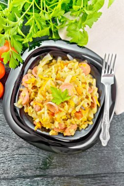 Siyah bir tabakta sosisli lahana güveci, peçete, domates, maydanoz ve çatal yukarıdan kara ahşap bir arka planda.