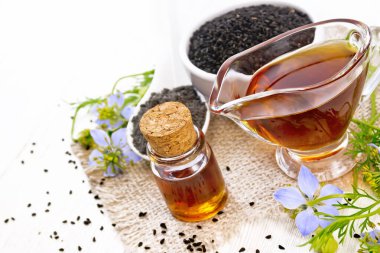 Nigella sativa yağ şişesi ve sos tekne, bir kaşık ve siyah kimyon un içinde çuval bezi, kalingi dalları mavi çiçekler ve beyaz ahşap arka planda yapraklar