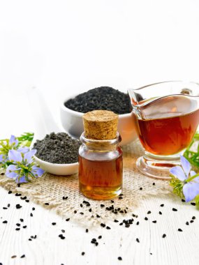 Nigella sativa yağı şişe ve sos kabında, kaşık içinde tohumlar çuval bezinde siyah kimyon unu, mavi çiçekli kalingi dalları ve tahta arka planda yapraklar.