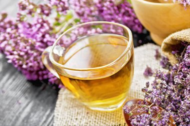 Çuval peçetede cam bir fincanda Oregano bitkisel çayı, çuval ve kaşıkta kurutulmuş marjoram çiçekleri, havanda taze çiçekler ve koyu ahşap tahtaya karşı masa.