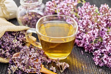 Cam bir fincanda Oregano bitkisel çayı, taze çiçekler, poşette kurutulmuş marjoram çiçekleri, koyu ahşap arka planda kavanoz ve kaşık