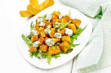 Fırında balkabağı salatası, tuzlu peynirli roka, ballı, hardallı, sarımsaklı ve sebze yağlı bir tabak, peçete ve çatallı beyaz tahta arka plan.