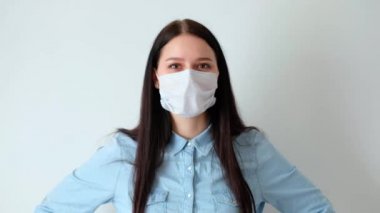 Genç ve gülümseyen bir kadına yakından bakın, uzun kahverengi saçlı bir kız tıbbi koruyucu bir maske takıyor, iç çekiyor, kameraya bakıyor, hijyen mantarı 19, koronavirüs, beyaz bir arka planda.