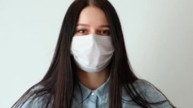 Genç ve gülümseyen bir kadına yakından bakın, uzun kahverengi saçlı bir kız tıbbi koruyucu bir maske takıyor, iç çekiyor, kameraya bakıyor, hijyen mantarı 19, koronavirüs, beyaz bir arka planda.