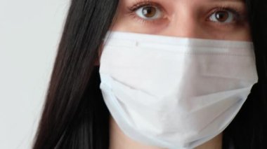 Uzun kahverengi saçlı, tıbbi koruyucu maske takmış genç bir kızın yakın plan fotoğrafı. Kameraya bakıyor. Hijyen mantarı, Coronavirus, beyaz arka plan çerçevesinde kayıyor.