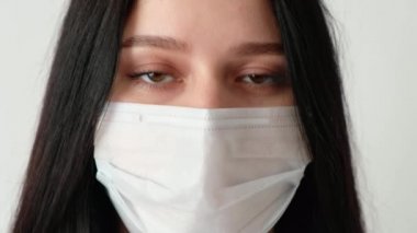 Uzun kahverengi saçlı, tıbbi koruma maskesi takmış genç bir kızın yakın plan fotoğrafı. Kameraya bakıyor. Hijyen mantarı 19, koronavirüs, beyaz bir arka plan.,