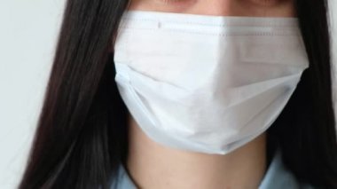 Uzun kahverengi saçlı, tıbbi koruyucu maske takmış genç bir kızın yakın plan fotoğrafı. Kameraya bakıyor. Hijyen mantarı, Coronavirus, beyaz arka plan çerçevesinde kayıyor.