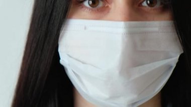 Uzun kahverengi saçlı, tıbbi koruyucu maske takmış genç bir kızın yakın plan fotoğrafı. Kameraya bakıyor. Hijyen mantarı, Coronavirus, beyaz arka plan çerçevesinde kayıyor.