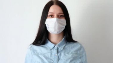 Genç ve gülümseyen bir kadına yakından bakın, uzun kahverengi saçlı bir kız tıbbi koruyucu bir maske takıyor, iç çekiyor, kameraya bakıyor, hijyen mantarı 19, koronavirüs, beyaz bir arka planda.