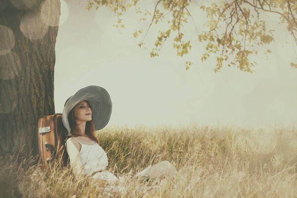 Beautiful woman in hat sitting near tree at countryside. Photo in old image color style