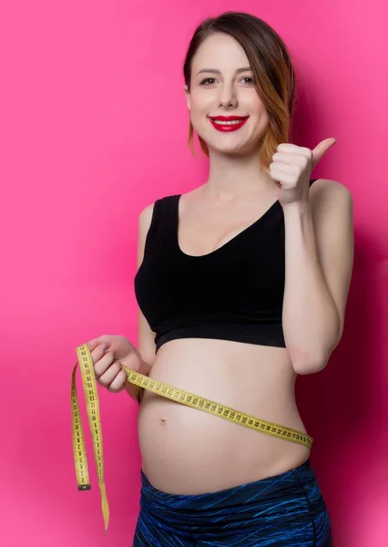 ピンクの背景に巻尺とフィットネスの服の若い妊婦 — ストック写真