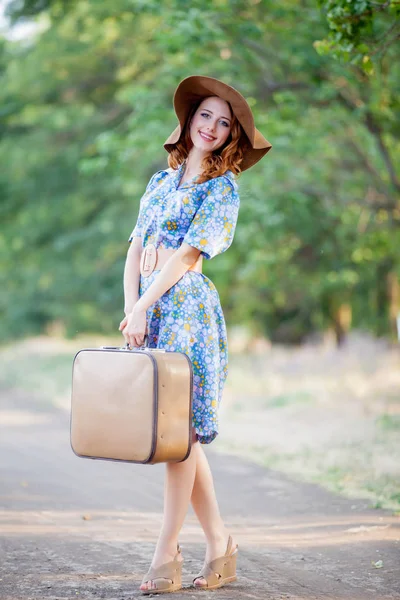 屋外の田園地帯では 道路上に立っているスーツケースと帽子でブルーのドレスの若い女性の肖像画 — ストック写真