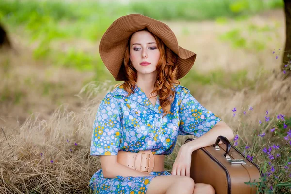 田舎屋外で草に座ってスーツケースと帽子でブルーのドレスの若い女性の肖像画 — ストック写真