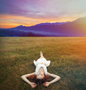 Young redhead girl lying down at green grass meadow with sunrise over mountines in summer time clipart