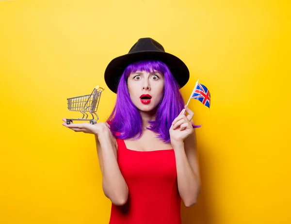 ショッピング カートと黄色の背景に手で英国旗紫髪型と若いスタイル内気な少女の肖像画 — ストック写真