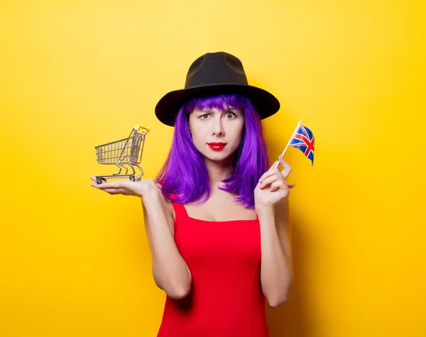 ショッピング カートと黄色の背景に手で英国旗紫髪型と若いスタイル内気な少女の肖像画 — ストック写真