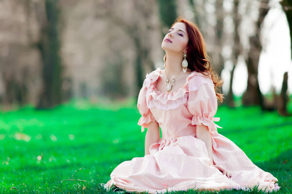 Portrait of a young redheadd girl in victorian style dress sitting on grass in springtime