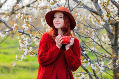 Kızıl saçlı kız kırmızı elbise ve şapka kiraz çiçeği bahçede poz kahve holding. 