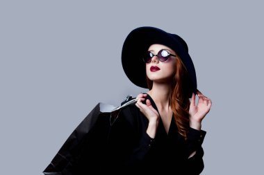 Güneş gözlüğü ve şapka gri arka plan üzerinde alışveriş çantası ayakta tutan genç zarif kadın 