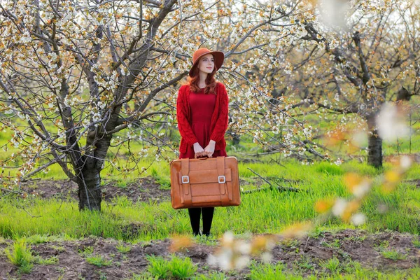 桜の花の庭 フルレングスでスーツケースと赤い服と帽子で赤毛の女の子 — ストック写真