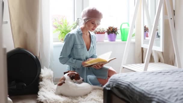 年轻的女孩与粉红色的头发和贝雷帽阅读书与狗在家里 时尚风格的服装 — 图库视频影像
