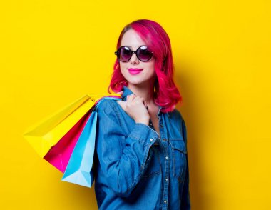 Genç saç kız güneş gözlüğü ve renkli bir alışveriş torbaları tutan mavi gömlek pembe. Portre izole Sarı zemin üzerine