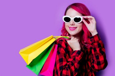Genç saç kız alışveriş torbaları ve güneş gözlüğü ile kırmızı ekose gömlekli pink. Mor arka plan üzerinde izole portre