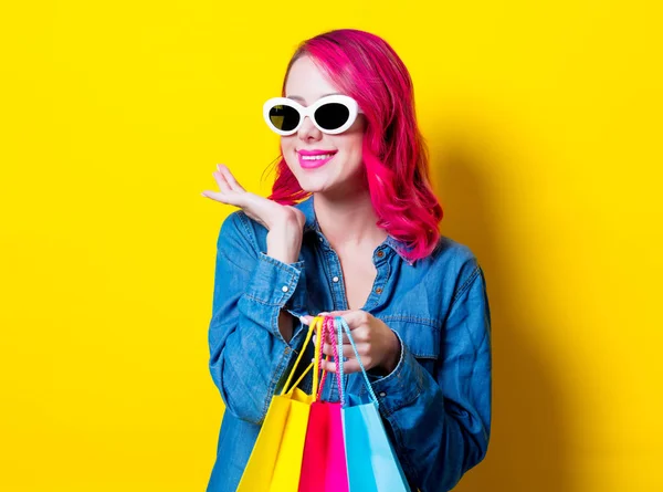 若いピンクのサングラスや色の買い物袋を保持している青いシャツの髪の少女 分離された黄色の背景の肖像画 — ストック写真